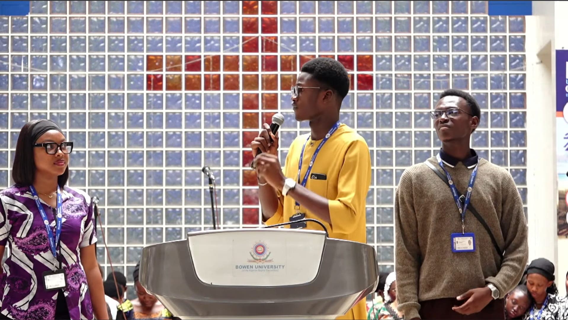 Abdul-Salam speaking at the Bowen University podium alongside two co-hosts